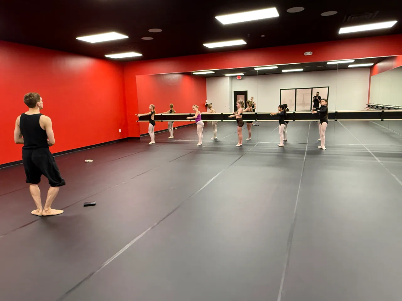 Young female dancers in leotards practice at the ballet barre under the instruction of Samuel Johnson in a studio with red walls.
