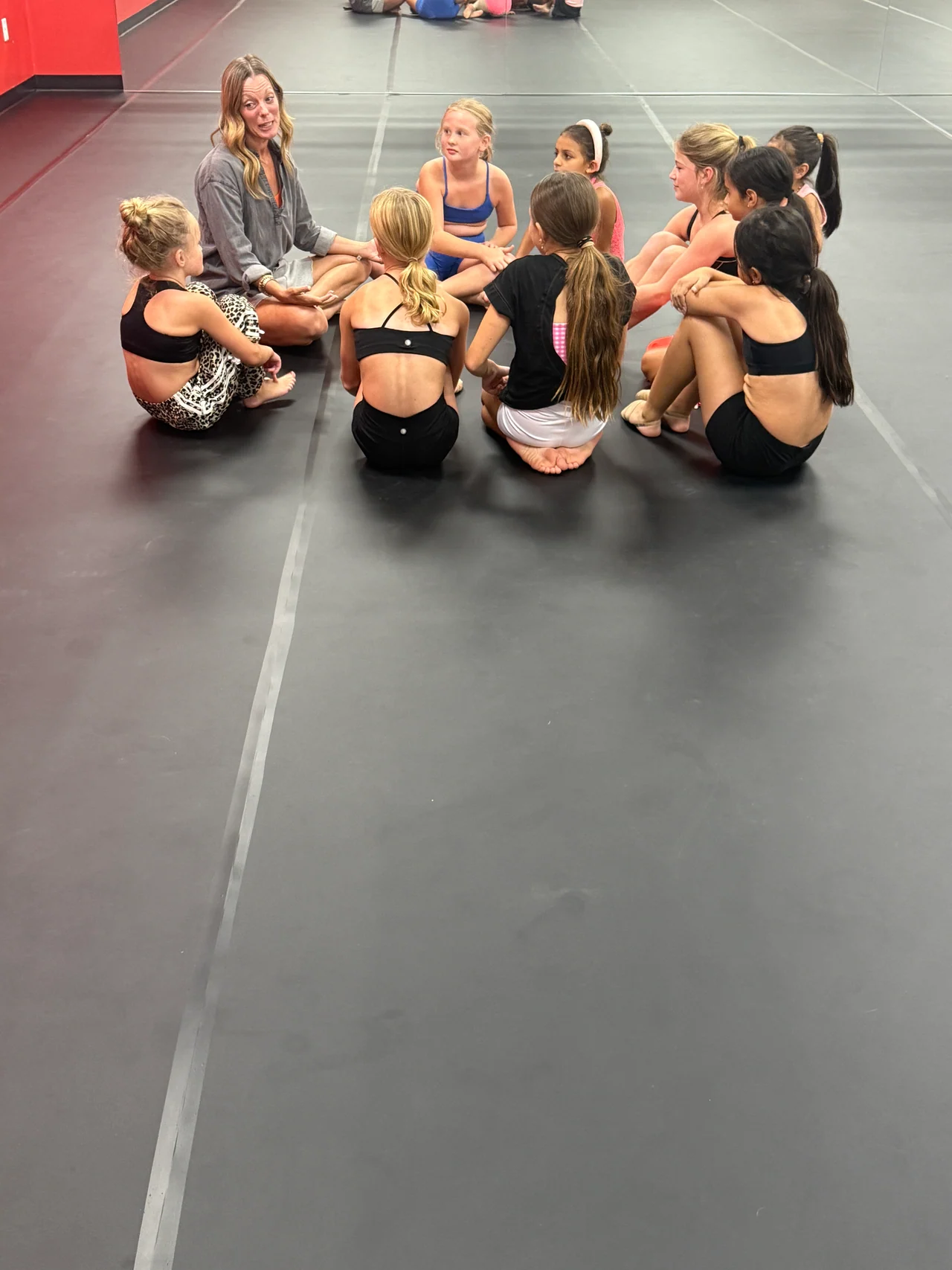 A group of young dancers and an instructor sitting together on a grey dance floor in the studio.